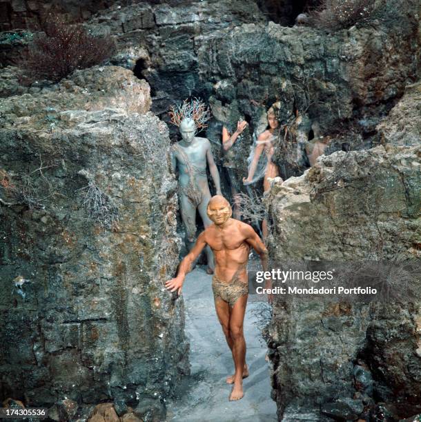Bare chested athletic man with a mask stands on a rocky scenery with other characters playing in Barbarella; all of them have fancy and coloured...