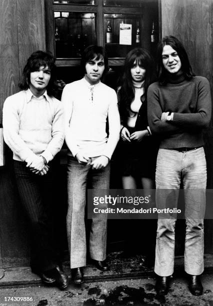 The four members of the Shocking Blue standing beside each other on a street, smiling at the camera, just near the window of a restaurant showing...