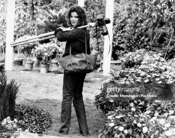 Italian television presenter and actress Gabriella Farinon walking in a garden with a camera with tripod on her shoulder. Rome, 1970s.