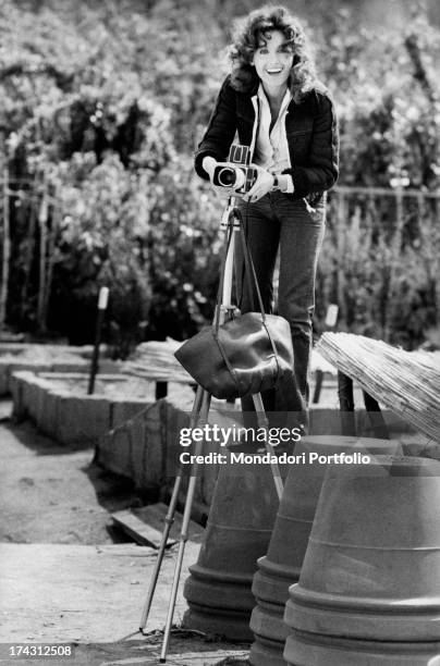 Italian television presenter and actress Gabriella Farinon, standing on a flowerpot, fixing a camera on a tripod. Rome, 1970s.