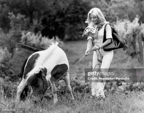 Italian television presenter and actress Gabriella Farinon in a field pulling a rope on which a pony is fastened. 1970s.