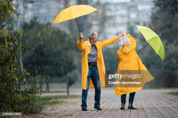 happy senior couple in raincoats dancing on rain in the park. - people rain happy stockfoto's en -beelden