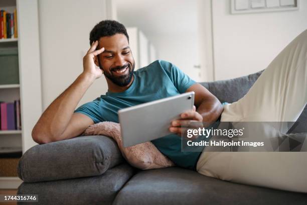 single man lying on sofa and binge-watching entire tv show on tablet. man watching a tv series on a streaming platform alone at home. enjoying the single life as young man. - serviço de distribuição digital contínua imagens e fotografias de stock