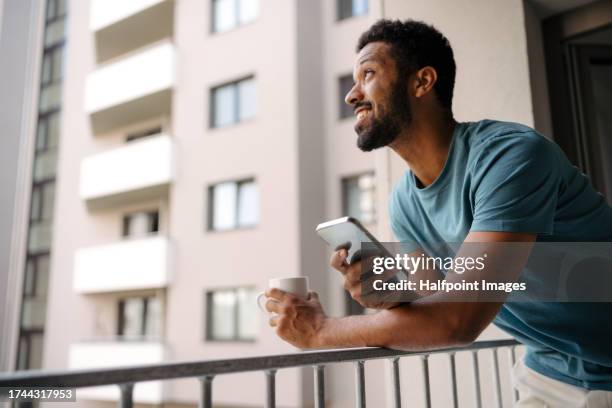 single man drinking first morning cup of coffee at home while scrolling on smartphone. standing on the balcony, leaning on railing and looking at the surroundings of his apartment building. - bachelor apartment stock pictures, royalty-free photos & images
