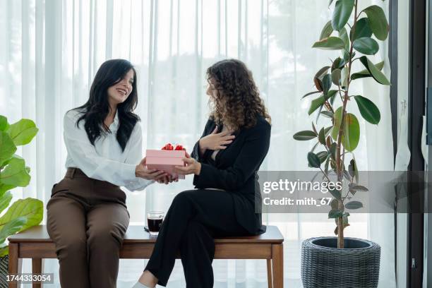 young adult asian female officer giving a new year gift to her caucasian close friend coworker at workplace, two female colleagues spending free time together and gift giving for thanking her about mental supporting in an office - exchanging gifts stock pictures, royalty-free photos & images