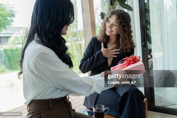 young adult asian woman giving christmas gift to her caucasian friend while sitting on a wooden bench at a cozy cafe and her friend looking happy, cheerful two women meeting with glad at cafe and talking about each other life - exchanging gifts stock pictures, royalty-free photos & images