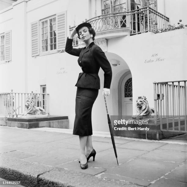 Secretary Janet Jones, undergoing training for a career in modelling, wearing a fitted suit with belted waist and pencil skirt outside a house in...