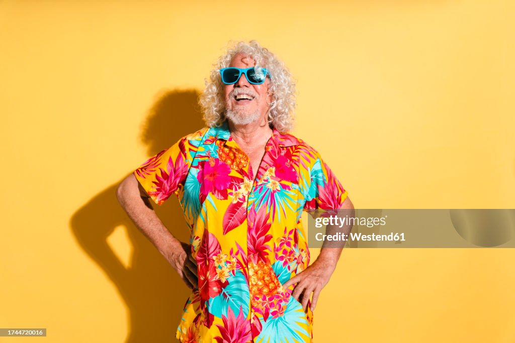 Happy senior man wearing sunglasses and standing with hands on hips against yellow background