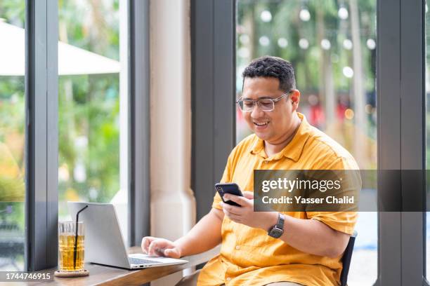 asian man using laptop at a cafe and looking at smartphone - indonesische etniciteit stockfoto's en -beelden