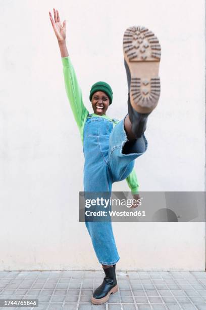 cheerful woman with leg-up and arm raised standing in front of white wall - schuhsohle stock-fotos und bilder