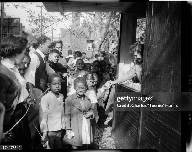 Small crowd of women, men, and children, including boy wearing foil crown, gathered in front of woman in booth handing out Sealtest ice cream cups,...