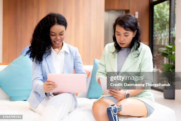 young businesswoman physical disability sitting on sofa looking digital tablet and laptop computer with partnership and talking together in office, business woman meeting and discussion with coworker. - small office lobby stock pictures, royalty-free photos & images