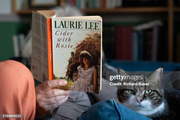 Family cat sits next to a woman who sits in her living room and reads a copy of Laurie Lee's seminal memoir, 'Cider With Rosie', on 20th October...