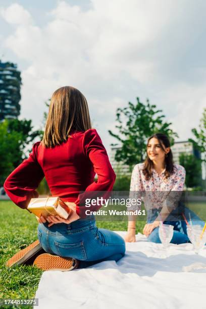 woman hiding gift box behind back in front of friend under cloudy sky - mani dietro la schiena foto e immagini stock