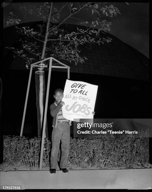 Boy holding sign inscribed 'Give to all, all at once, jobs' standing in front of Civic Arena, Lower Hill District, Pittsburgh, Pennsylvania, c. 1961