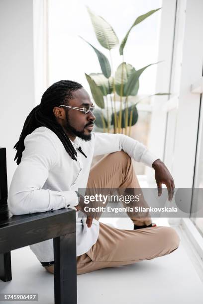 close-up portrait of pensive young african man sitting near the armchair at cozy living room. - portrait of pensive young businessman wearing glasses stock pictures, royalty-free photos & images