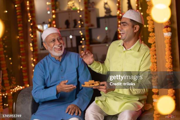 son offering sweet to father at illuminated home during eid-ul-fitr - indian muslims celebrate eid ul fitr stockfoto's en -beelden