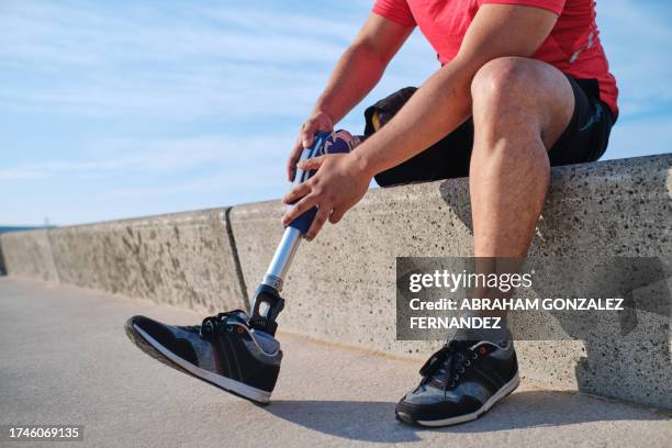 unrecognizable young man with one leg adjusting his prosthetic leg sitting on a retaining wall near the beach - prosthetic leg stock pictures, royalty-free photos & images