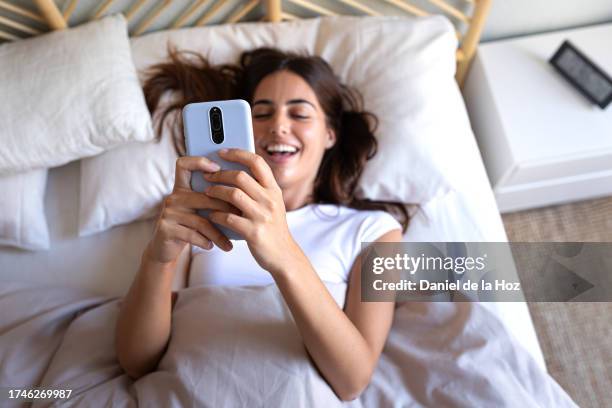happy young woman using mobile phone in bed. smiling female looking mobile phone in the morning after waking up. selective focus. - namoro pela internet imagens e fotografias de stock