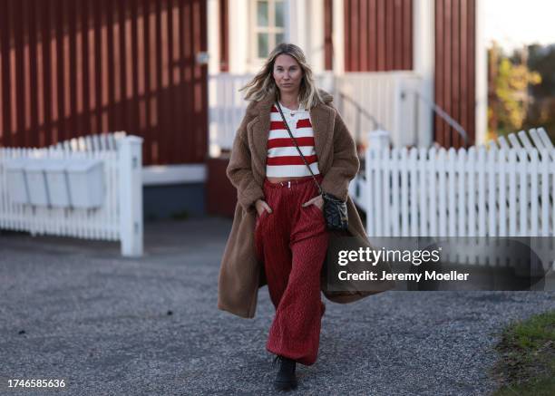 Karin Teigl seen wearing a red/white striped knit pullover, red print pattern wide pants, black leather crossbody bag, brown fluffy long teddy coat...