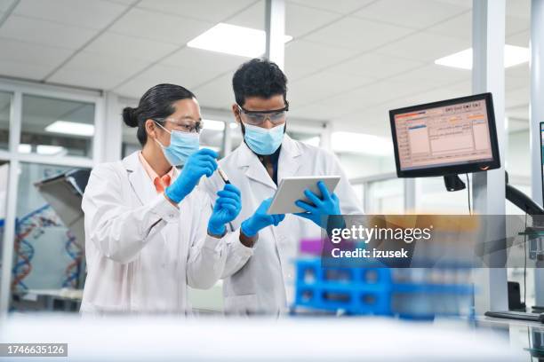 female and male biochemists wearing protective face masks while discussing over digital tablet at laboratory - clinical trials stock pictures, royalty-free photos & images