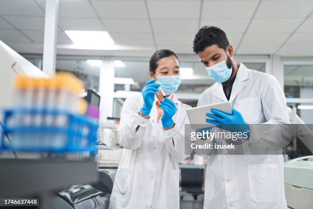 male and female scientists wearing protective face masks while discussing over digital tablet at laboratory - scientist at work stock pictures, royalty-free photos & images