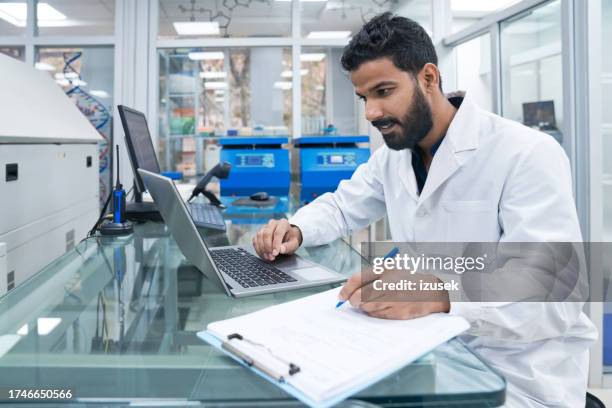 young male biochemist using laptop while writing on clipboard at laboratory - clean suit stock pictures, royalty-free photos & images