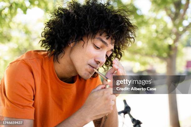a young man lighting a marijuana cigarette in the park - smoking weed stock pictures, royalty-free photos & images
