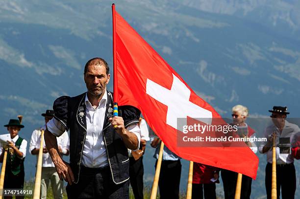 Flag thrower looks on as alphorn players prepare to perform on July 28, 2013 in Nendaz, Switzerland. About 150 alphorn blowers performed together on...