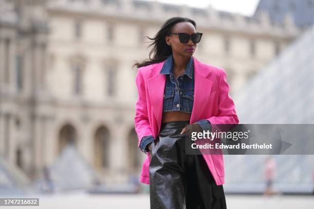 Emilie Joseph wears sunglasses, a blue cropped denim shirt, a neon pink oversized blazer jacket, a black gathered shiny leather midi skirt, during a...