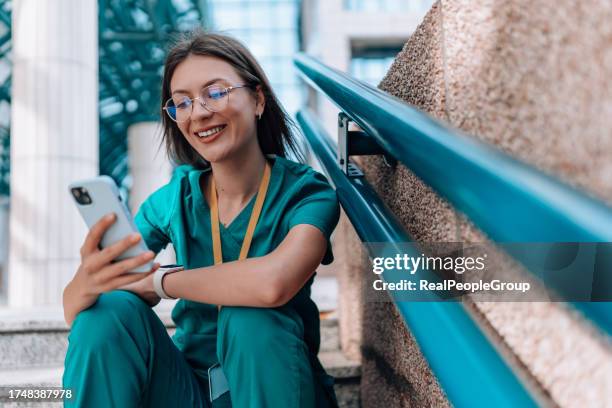 junge krankenschwester sitzt in ihren kitteln auf der treppe, während sie fröhlich lächelt und durch die sozialen medien scrollt - medizinerkleidung stock-fotos und bilder