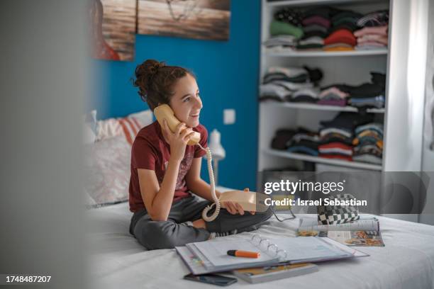 teenage girl sitting on her bed, holding an antique landline phone to talk to her friends while taking a break from her schoolwork. - telefone fixo imagens e fotografias de stock