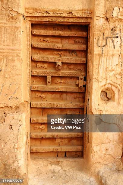 Heavy woodeen door with intricate geometric carvings features a large metal knocker and is held together by wooden straps and metal rivets, in...
