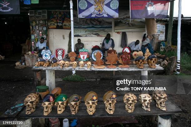 Artisans sit behind a display of some of their carvings that depict human skulls in beret's bearing military insignias of various British Army...