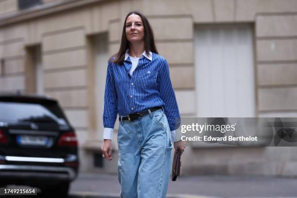 Alba Garavito Torre wears a blue and white striped shirt, a leather belt, blue flared denim jeans / pants, a brown leather clutch, during a street...