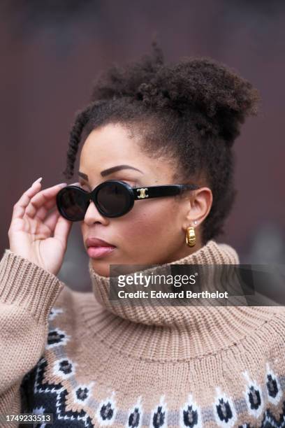 Ellie Delphine wears earrings, Celine sunglasses, a beige / pale brown wool turtleneck pullover with black printed geometric patterns from Khaite,...