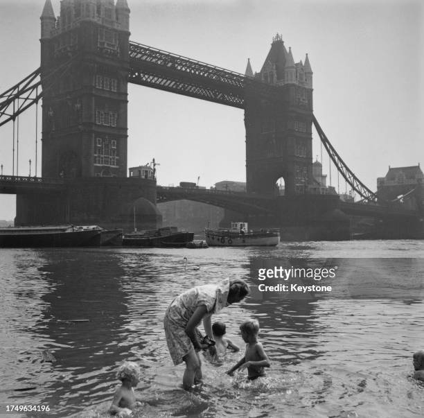 Woman paddles as children play in the waters of the River Thames off Tower Beach on the north bank of the river, Tower Bridge rising beyond, London,...