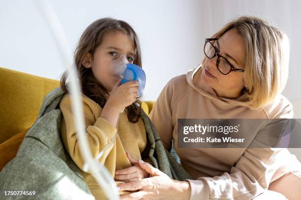 sick and tired little girl in mask with inhaler makes inhalations while sitting on the couch with her middle aged mother - equipamento-respiratório - fotografias e filmes do acervo