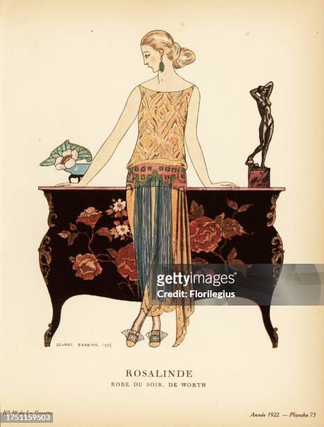 Woman in evening dress of gold lace with fringed belt leaning against a chinoiserie sideboard. Rosalinde. C’est une robe du soir, de Worth, en...