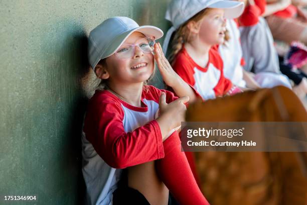 adorable elementary age girl adjusts her glasses while sitting in dugout with little league baseball team - baseball huddle stock pictures, royalty-free photos & images