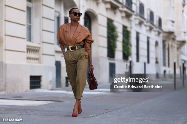 Emilie Joseph wears golden earrings, a vintage camel leather jacket worn as a top, high-waisted 7/8 paper bag pants; a black leather belt embellished...