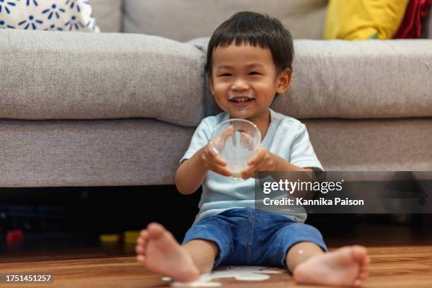 adorable asian little boy spilled milk all over the floor while drinking a glass of milk in living room at home. - knoeien stockfoto's en -beelden