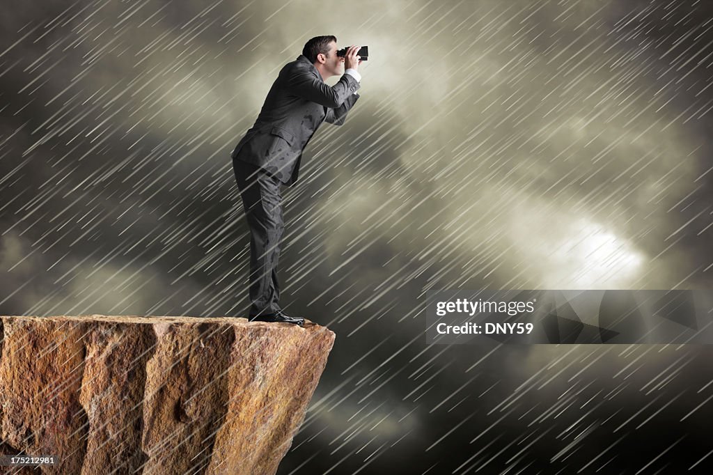 Businessman standing on cliff in driving rain looking through binoculars