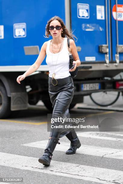 Guest wears sunglasses, one earring, necklaces, a white top, a large black leather belt, a black leather midi skirt, black leather biker boots,...