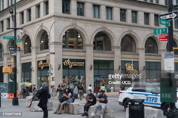 Wide view of the entrance and SE corner of Wegmans store as seen from Astor Place Plaza, New York, New York. More than 30 people, engaged in a...