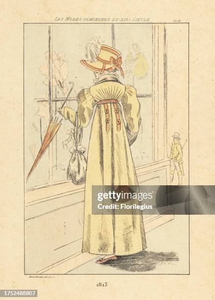 Fashionable woman window-shopping for bonnets outside a milliners, Paris, 1813. She wears a bonnet, high-waisted dress with full sleeves, holding a...