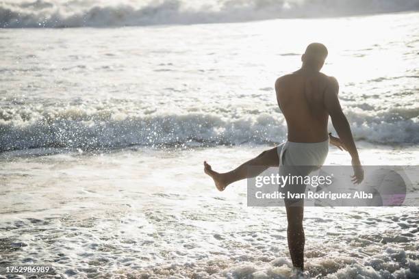 african american boy enjoys kicking the sea water on the beach shore. - kicking sand stock pictures, royalty-free photos & images