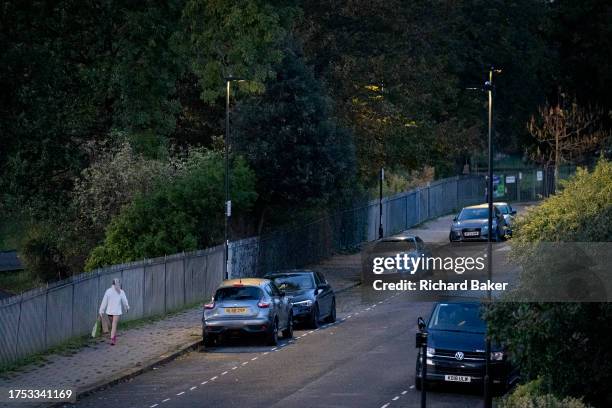 Seen from a high perspective, a lone woman walks at dusk past parked cars on a well-lit residential street in Lambeth, south London, on 27th October...