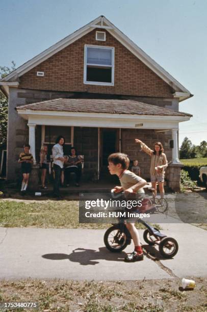 Kevin Watkins is seen in front of the new family home, riding a tricycle as his family and neighborhood children congregate on the porch ca. 1975.