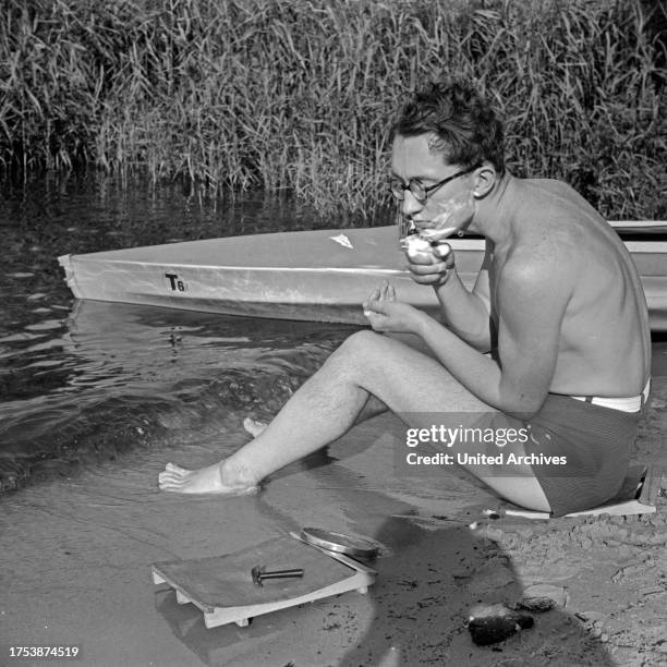 Young man shaving at the shore of a lake, beside a Klepper folding boat, Germany 1930s.